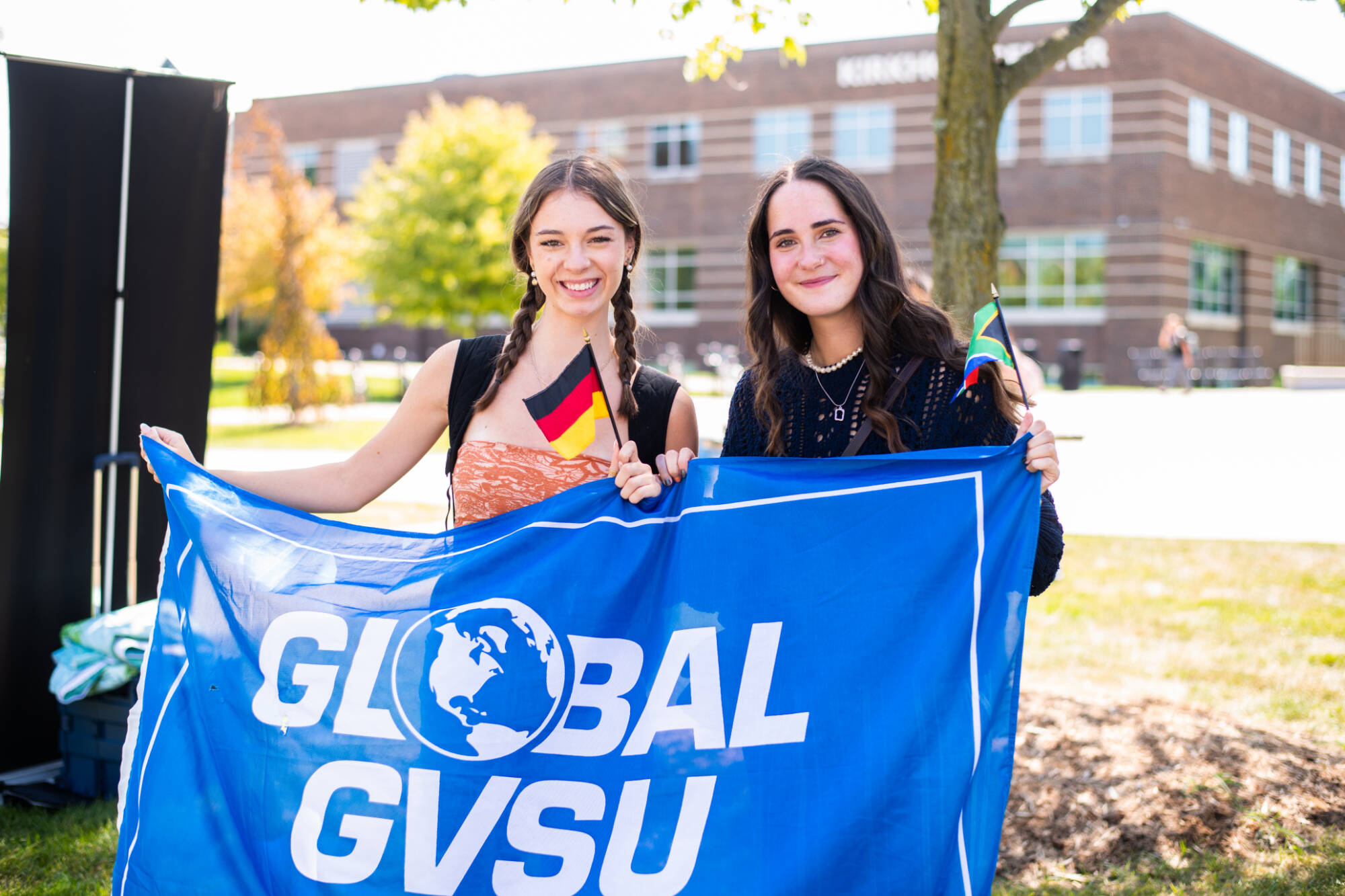 two students standing behind a banner that reads "global GVSU" while waving tiny flags for non US countries